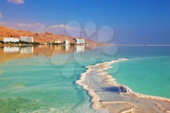 Israeli coast of the Dead Sea. Path from the salt winds picturesquely of salt water. Hotels on the bank are reflected in smooth water