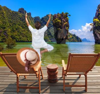 Exotic vacation in Thailand. Two beach chairs and an elegant hat on one. Woman performs yoga pose Tree on the Bay
