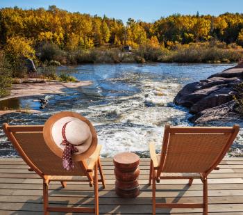 Two wooden chairs and hat on the shore of Winnipeg River. Foam water rapids on the smooth stones. Old Pinawa Dam Park. The concept of ecological and recreational tourism