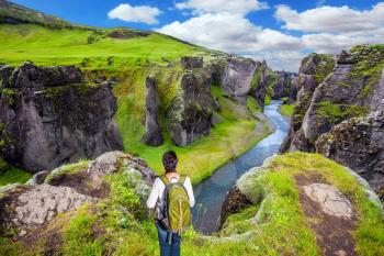  Green Tundra in summer. The concept of active northern tourism. The elderly woman -  tourist with big backpack admiring the magnificent scenery. The striking canyon in Iceland