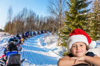 Soon Christmas. Concept of active winter tourism. Handsome boy in a red Santa Claus hat. Tourist train from snowmobiles moves along the ice of a frozen river