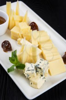 Cheese plate on a dark background table. Many kinds of cheese with sauce and greens on a white plate, closeup shot