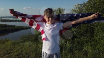 Blonde boy waving national USA flag outdoors over blue sky at the river bank.