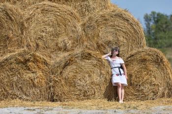 A brunette girl in a white dress and glasses, against a background of a large haystack from round bales.