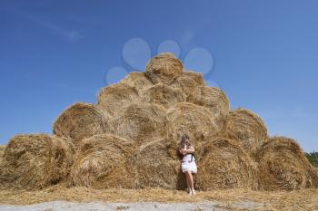 A young brunette girl next to a large haystack of round bales laid out in the form of a pyramid