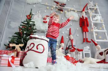 Little girl in a red sweater takes artificial snow from a bag and throws it against the background of the Christmas interior with gnomes, a ladder and garlands