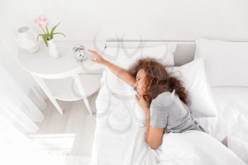 Morning of sleepy young African-American woman turning off alarm clock while lying in bed�