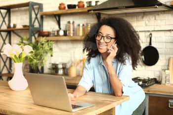 Female African-American freelancer talking by phone while working on laptop in kitchen�