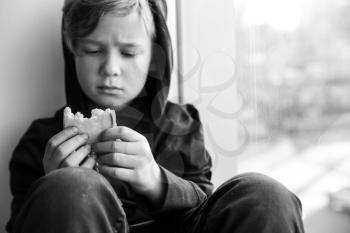 Homeless little boy with bread sitting on window sill indoors�