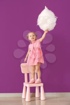 Cute little girl with cotton candy standing on chair against color wall�