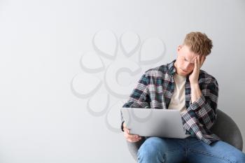 Tired young man with laptop sitting on chair against light background�