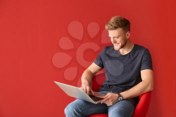 Handsome young man with laptop sitting on chair against color background�