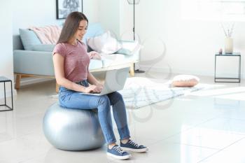 Young woman with laptop sitting on fitball at home�