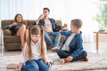 Sad little children sitting on floor while addicted parents drinking alcohol at home�