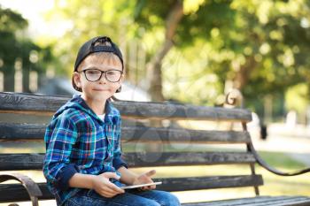Cute little boy with tablet computer sitting on bench in park�