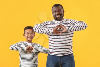 Portrait of African-American man and his little son making hearts with their hands on color background�