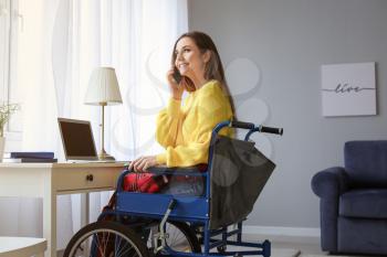 Handicapped young woman in wheelchair talking by phone while using laptop at home�