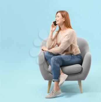Beautiful woman talking by phone while sitting in armchair against color background�
