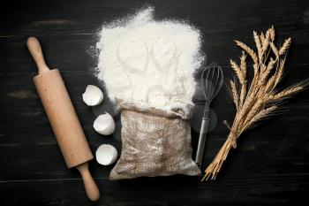 Bag with flour, utensils, egg shell and wheat spikelets on dark background�