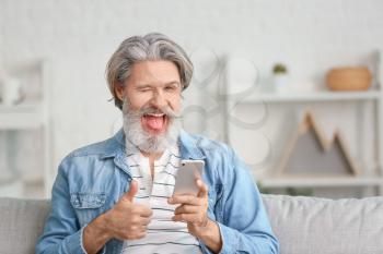 Happy elderly man with mobile phone showing thumb-up gesture at home�