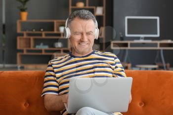 Mature man with laptop and headphones sitting on sofa at home�