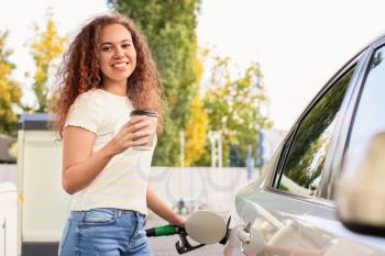 African-American woman drinking coffee while filling up car tank at gas station�