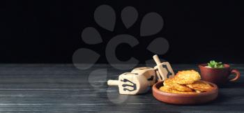 Dreidels and potato pancakes for Hanukkah on table against dark background with space for text�