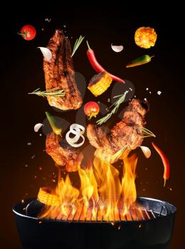 Falling tasty meat with vegetables and spices on barbecue grill with flame against dark background�