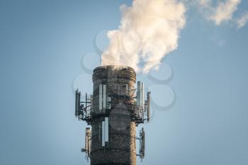 Top of high industrial chimney with multiple cell phone antennas, smoke comming out on clear blue sky background 