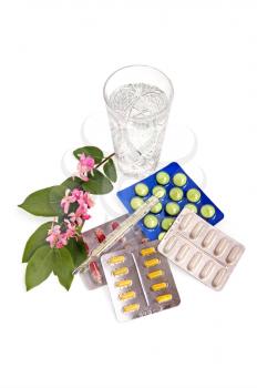 A set of tablets and capsules with a thermometer, the water in a crystal glass and a branch with pink flowers and green leaves on a white background