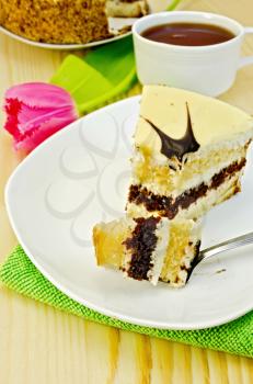 Sponge cake with cream and a fork on a plate, a cup of tea, pink tulip on a green napkin against a wooden board