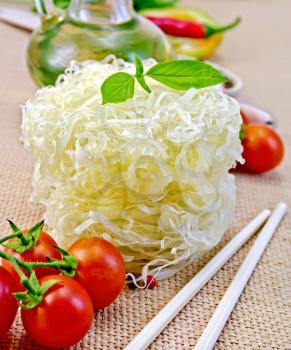 Rice noodles twisted, tomatoes, different pepper, oil in carafe, chopsticks on a background of sack cloth