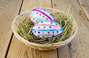 Two easter eggs, decorated with multicolored braid and sparkles in the hay and a wicker basket on a wooden boards background