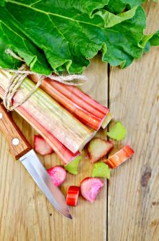 Bundle of stalks of rhubarb leaf and cut pieces of rhubarb, knife on background wooden plank