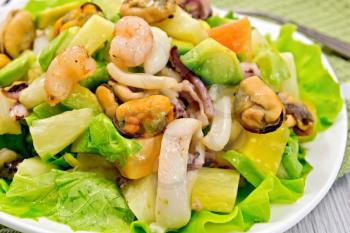Salad with shrimps, octopus, mussels and calamari with avocado, lettuce, pineapple on a plate, fork, napkin on the background light wooden boards