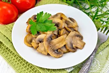 Champignons fried in a plate with parsley leaf, fork, ripe tomatoes on a green napkin on the background light wooden boards