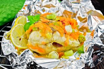 Pike with carrots, leek, basil and lemon slices in a foil on a metal grid, towel on a dark wooden board