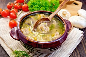 Soup with meatballs, noodles and mushrooms in a clay bowl with a spoon on a napkin, parsley, tomatoes and bread on a dark wooden board