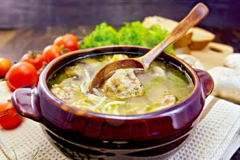 Soup with meatballs, noodles and mushrooms in a clay bowl with a spoon on a napkin, parsley, tomatoes and bread on a wooden boards background