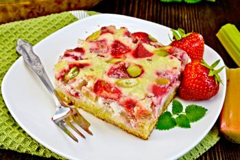 Piece of pie with strawberries, rhubarb and cream sauce, fork, strawberry, mint in white plate on a green towel, rhubarb stalks on a wooden boards background