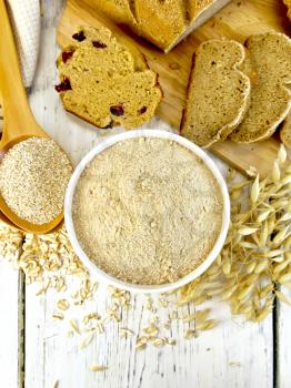 Oat flour in white bowl, oat bran in spoon, oatmeal and stems, bread and biscuits on a background of wooden boards on top