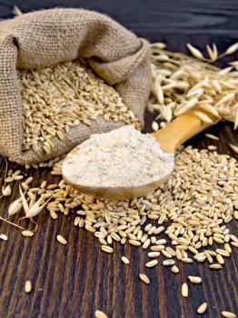 Oat flour in a wooden spoon, a bag of oats and oat stalks on a dark wooden board