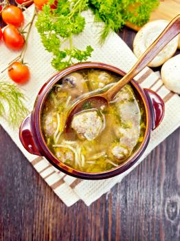 Soup with meatballs, noodles and champignon in a clay bowl with spoon on a napkin, parsley, mushrooms, tomatoes and bread on background a wooden board on top