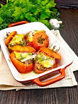 Tomatoes stuffed with meat and rice with cheese in a brazier on a towel, parsley, dill and garlic on a wooden board background