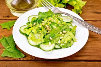 Salad from spinach, fresh cucumbers, rukkola salad, cedar nuts and spring onions, seasoned with vegetable oil and a fork on a plate on the background of a wooden board