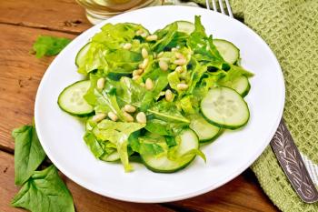 Salad of spinach, fresh cucumbers, rukkola salad, cedar nuts and green onions, seasoned with vegetable oil in a plate, napkin and fork on a plank background