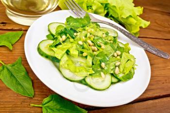 Salad from spinach, fresh cucumbers, rukkola salad, cedar nuts and spring onion seasoned with vegetable oil and spices with a fork on a plate on a wooden board background