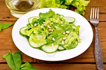 Salad from spinach, fresh cucumbers, rukkola salad, cedar nuts and spring onions, seasoned with vegetable oil on a plate, fork on a wooden plank background