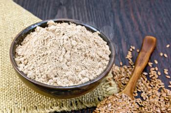 Flax flour in a bowl on napkin from burlap, seeds in a spoon and on table on the background of wooden board