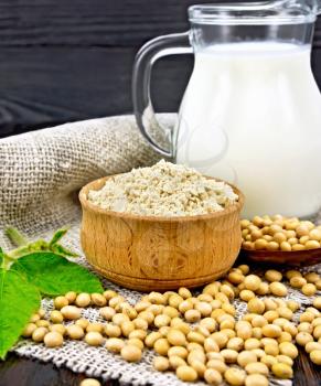 Soy flour in the bowl, soybeans in a spoon and on a napkin of burlap, milk in a jug, soya leaf against the background of a dark wooden board
